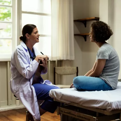 A young woman sits in a hospital room and talks about her health and wellness with her primary care doctor.