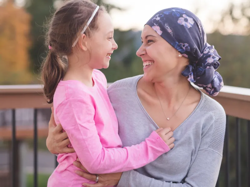 A mother and cancer survivor is sitting, with one arm around her daughter.