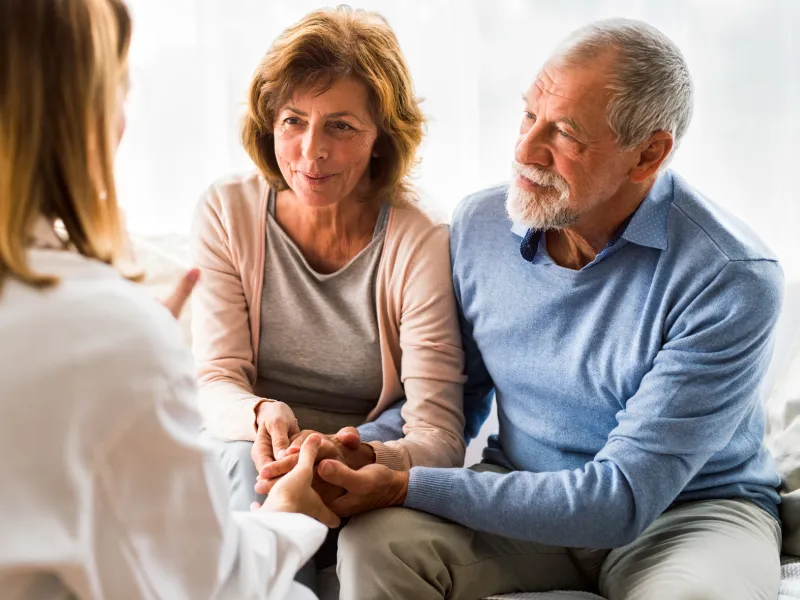 A senior couple, sitting down and holding hands, consulting a doctor at an appointment.