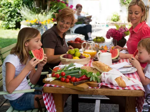 A family of four enjoys a summer picnic, outdoors, sitting at a picnic table enjoying summer foods.