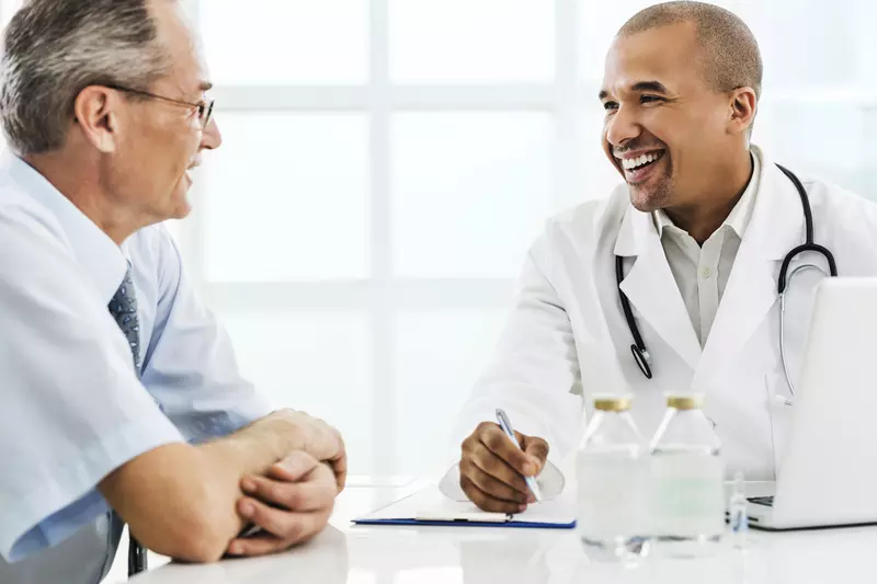 A senior man visits his male doctor for his annual screening. Both are seated at a table.