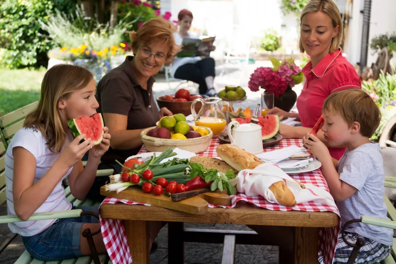 A family of four enjoys a summer picnic, outdoors, sitting at a picnic table enjoying summer foods.