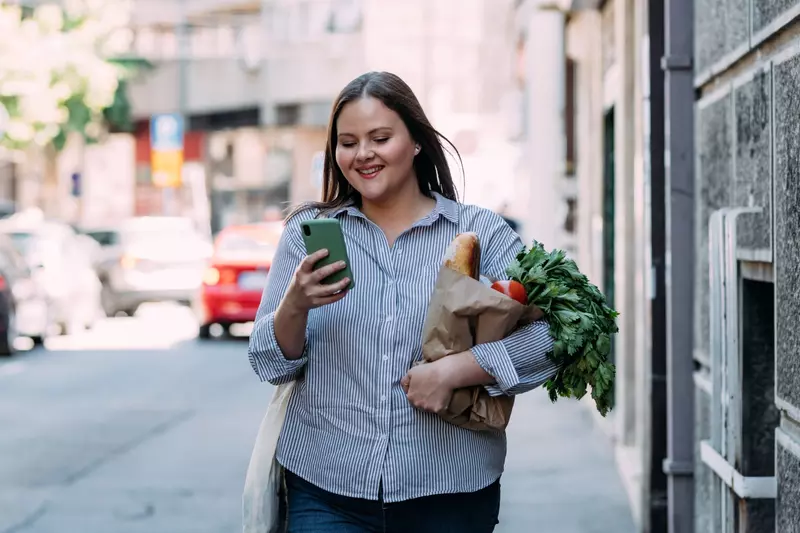 Smiling woman walking and looking at her phone while carrying a bag of groceries.