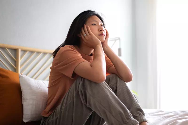 An Asian woman sits on a bed staring with a look of ennui.