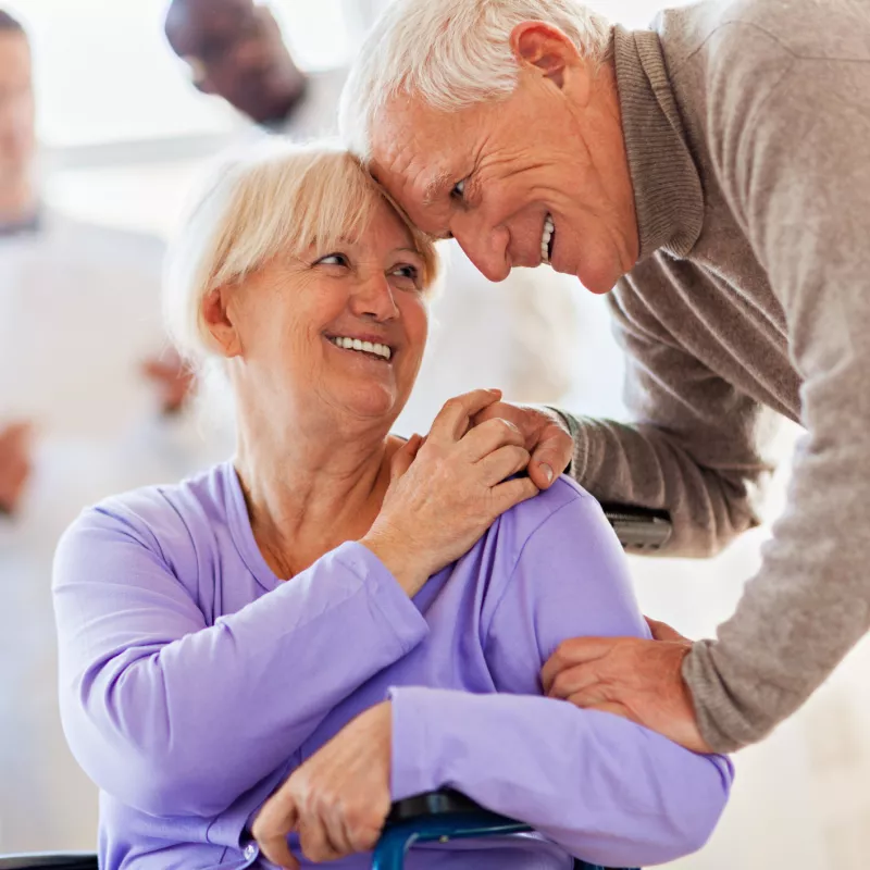 Elderly couple, the woman is sitting in a wheelchair and the man is bending down, his hands on her shoulders, comforting her.