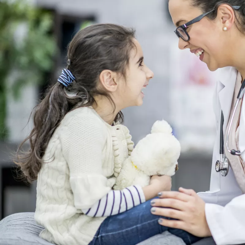 A child sitting on an exam table with her doctor, who has her hand on her knee and is smiling at her.