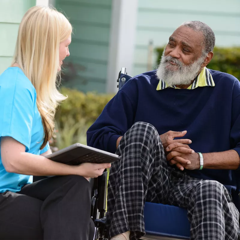 Elderly man in a wheelchair outside speaking to a nurse.