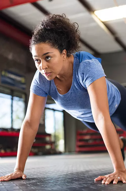 A young African American woman with a blue shirt performs push-ups at an open spaced gym.